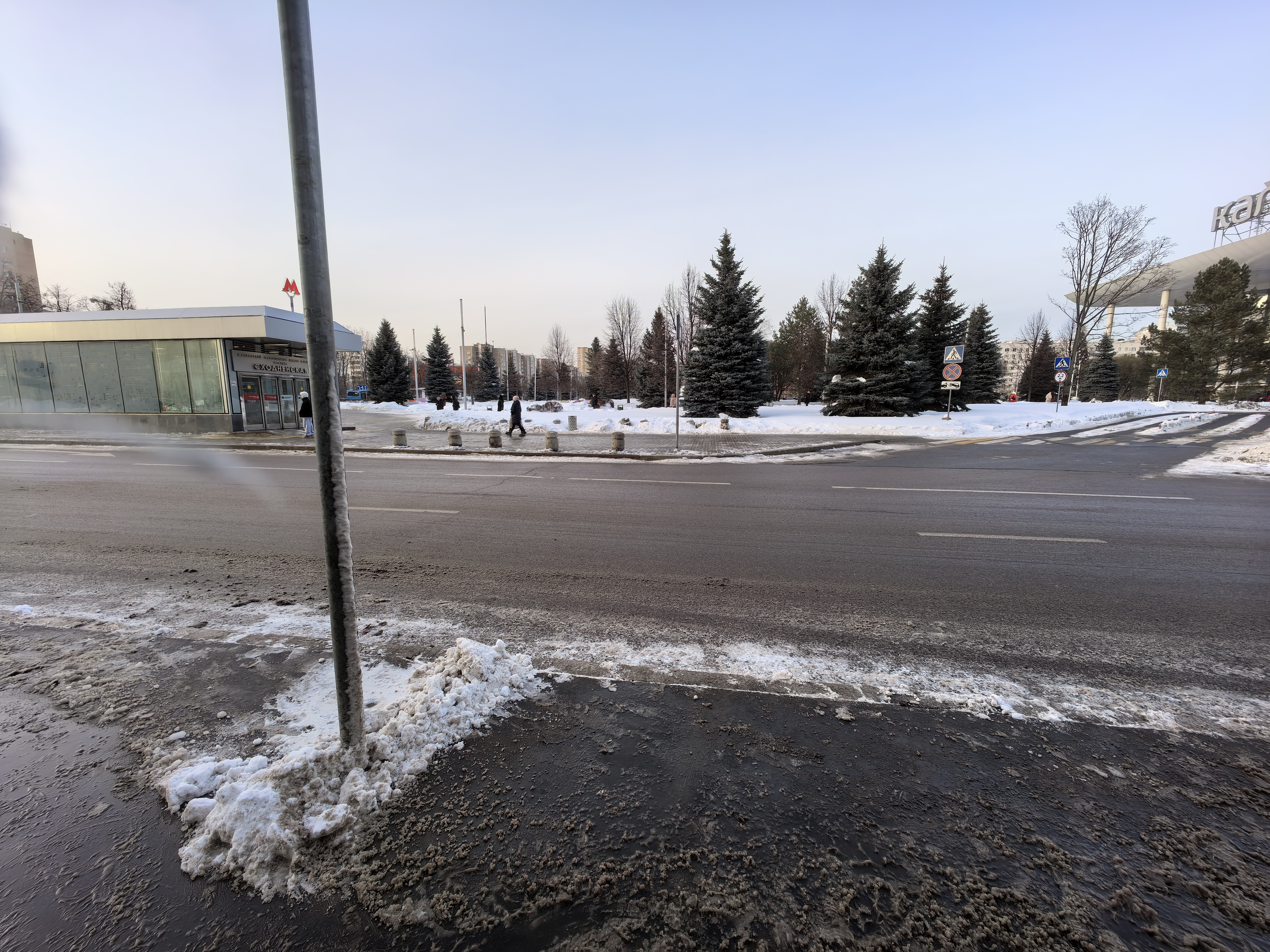 Широкая городская проезжая часть с пешеходным переходом, расположенная рядом с входом в метро и парковой зоной. Видны дорожная разметка, знаки перехода и ограждающие столбики.