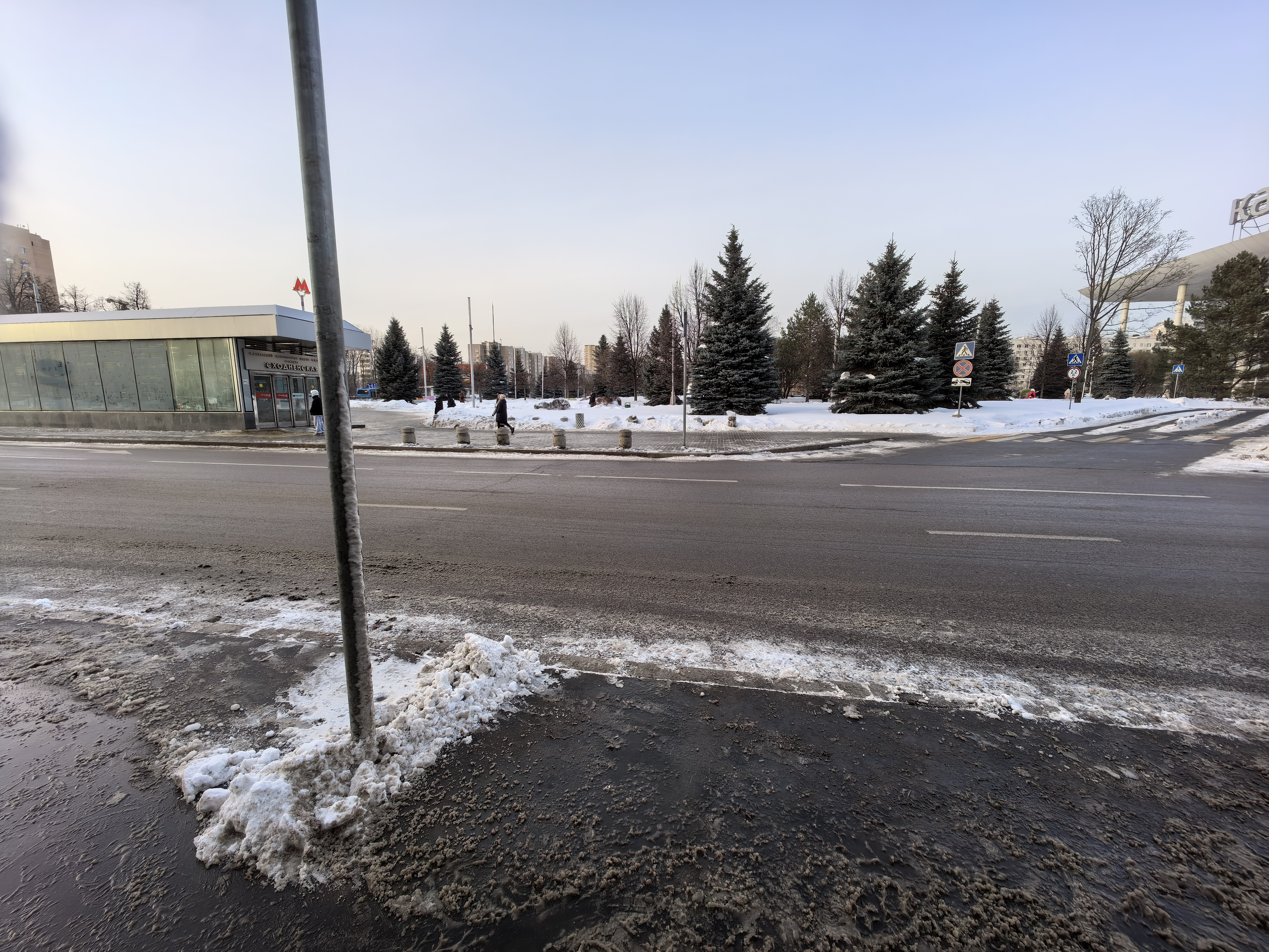 Участок городской дороги с пешеходным переходом, расположенный у входа в метро. На проезжей части видна разметка, тротуары есть, но покрыты снегом.